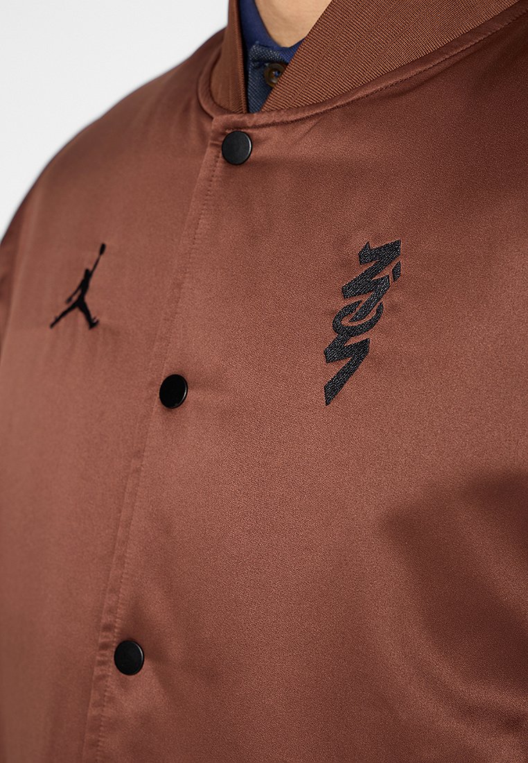 Giacca bomber in satin marrone con bottoni a pressione neri, dotata di un logo Jumpman nero e ricamo in testo stilizzato nero sul lato sinistro del petto.