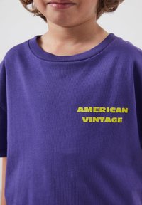 Enfant portant un t-shirt violet avec le texte jaune "AMERICAN VINTAGE" sur le côté gauche de la poitrine, cadré au niveau du cou et des épaules.
