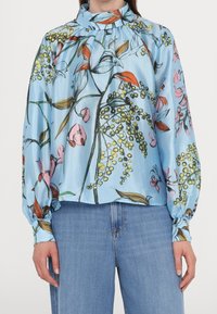 Donna che indossa una blusa a maniche lunghe di un azzurro chiaro con una stampa floreale e colletto alto, abbinata a jeans di denim blu.