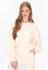 Creme, strikket sweater med strukturerede ærmer, blomster mønstre og ribbet kant. Kombination med lyse bukser, model står foran en hvid baggrund.