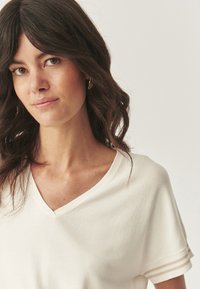 T-shirt a manica corta color beige chiaro con scollo a V, realizzato in un tessuto morbido. Presenta due sottili strisce vicino ai polsini e un fit rilassato.