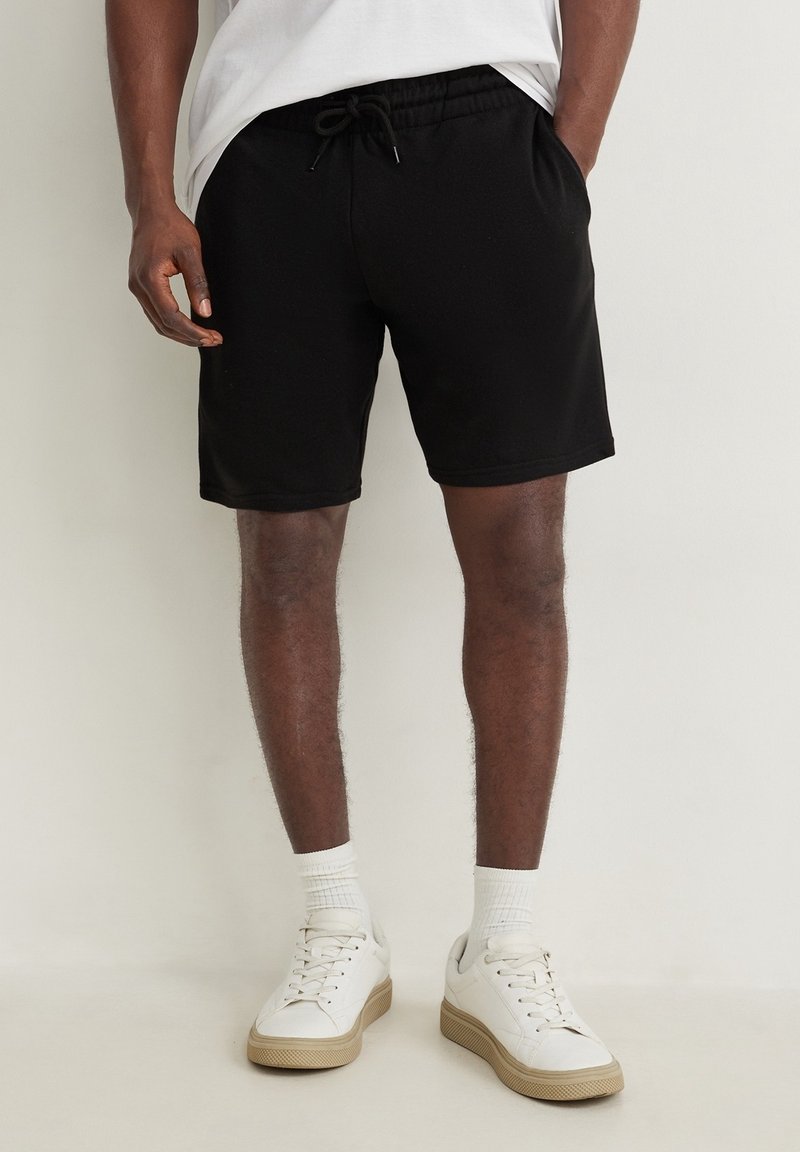 Homme portant un short noir à cordon, des chaussettes blanches montantes, des baskets blanches et une chemise blanche, les mains dans les poches, sur un fond uni.