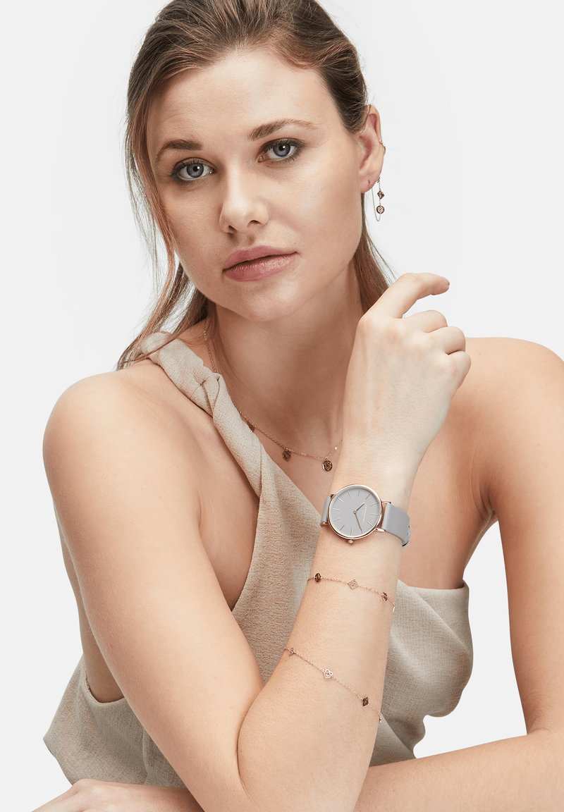 Montre grise à cadran rond et accents métalliques portée au poignet. Le modèle porte un haut en tissu beige et des bracelets en rose doré superposés.