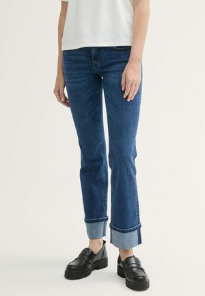 Jean en denim bleu avec une coupe droite, présentant des ourlets retroussés et un design classique à cinq poches. Associé à des chaussures noires à enfiler.