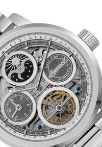 Montre en acier inoxydable avec un cadran ajouré complexe, deux fuseaux horaires, des aiguilles en ton argent et des accents dorés sur les engrenages.
