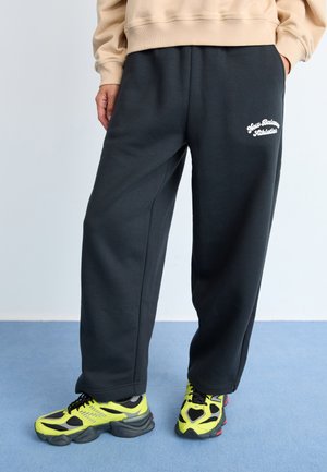 Zwarte joggingbroek met een relaxte pasvorm, voorzien van een wit geborduurd logo. In combinatie met opvallende gele en zwarte sportschoenen.