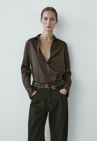 Camicia a righe marroni con bottoni e scollo profondo, abbinata a pantaloni neri a vita alta e una cintura intrecciata in pelle.