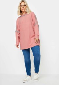 Vaate pitkähihainen vaaleanpunainen huppari, jossa on sivu- ja yksityiskohtia, yhdistettynä sinisiin kapeisiin farkkuihin ja valkoisiin lenkkareihin. Suora pinta, rento leikkaus ja pyöreä kaula-aukko.