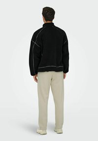 Pull en polaire noir avec un col montant, arborant des coutures blanches contrastantes. Porté par-dessus un pantalon beige clair et associé à des chaussures claires.