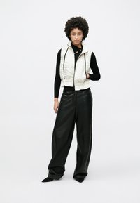 Weiße gesteppten Weste mit dekorativen Nähten, schwarzes Langarmshirt und lockere schwarze Lederhose. Das Model steht vor einem schlichten Hintergrund.