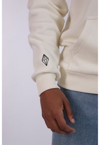 Sudadera color crema con puños y cintura acanalados. Cuenta con un logotipo geométrico bordado en negro en la manga. Jeans de mezclilla visibles por debajo.