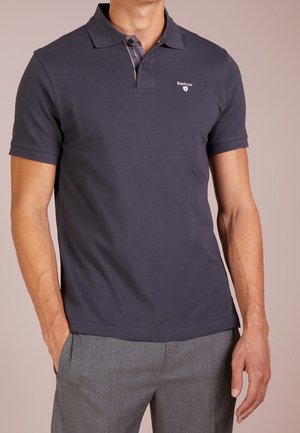 Mann trägt ein dunkelgraues Barbour-Poloshirt mit kurzen Ärmeln und graue Hose, steht mit einer Hand in der Tasche.