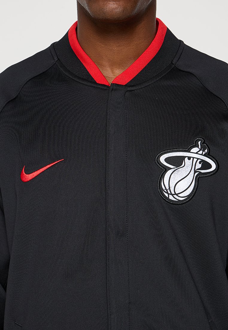 Giacca nera con un colletto e dettagli rossi. Presenta un logo Nike rosso e un logo Miami Heat bianco sul lato sinistro del petto. Texture liscia.