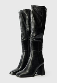 Juodi iki kelių siekiantys batai pagaminti iš lygios odos, su kvadratiniu kulnu ir šoniniu užtrauktuku, turintys elegantišką ir struktūruotą dizainą.