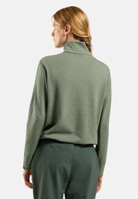 Pullover verde con collo alto, realizzato in materiale testurizzato. Maniche lunghe con spalle scese, indossato con pantaloni verdi abbinati.