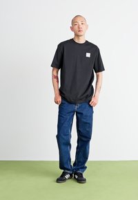 T-shirt noir à manches courtes avec une poche poitrine et un jean en denim bleu, associé à des baskets noires avec des rayures blanches ; pose debout.