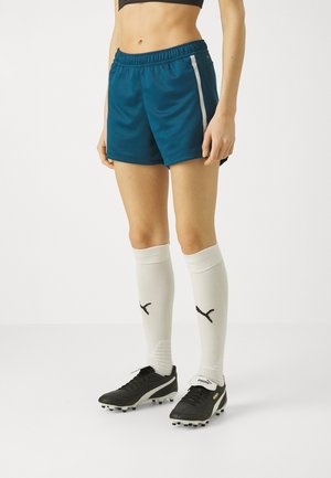Person iført mørk teal sportsshorts med en hvid lynlås, hvide knæhøje Puma-sokker og sorte Puma-fodboldstøvler på hvid baggrund.