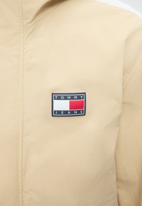 Μπεζ σακάκι με ορθογώνιο λογότυπο Tommy Jeans με λευκά και κόκκινα χρωματιστά μπλοκ στην περιοχή του στήθους.