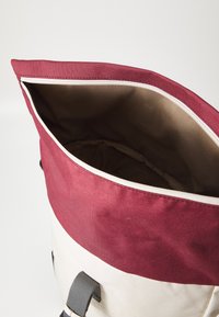 Geantă din pânză cu un vârf burgund, un corp crem și un interior bej. Dispune de un închizător cu fermoar și curele din țesătură gri.