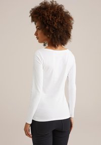 Frau mit lockigem Haar, die ein weißes Langarmshirt und schwarze Hosen trägt, von hinten gegen einen einfarbigen beigen Hintergrund betrachtet.