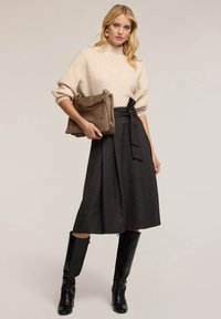 Maglione a coste beige, gonna grigia plissettata con nodo laterale e stivali neri in pelle al ginocchio. La modella tiene una borsa strutturata marrone.