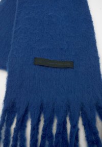 Blå tørklæde lavet af blødt, tekstureret materiale med frynseendelser. Har et sort mærke med "KLASSISK. MODERNE. DIT." prægning.