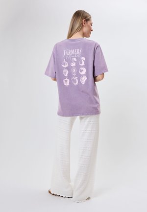 Kobieta z długimi włosami stoi tyłem, ubrana w lawendową koszulkę z białymi ilustracjami owoców i kwiatów oraz białe, wzorzyste spodnie z szerokimi nogawkami.