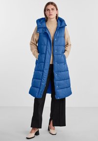 Mėlyna pūkinė liemenė su kapišonu, su siūtu raštu, užtrauktuku ir spaustukų akcentais. Dėvima ant smėlio spalvos golfuko ir juodų platučių kelnių.