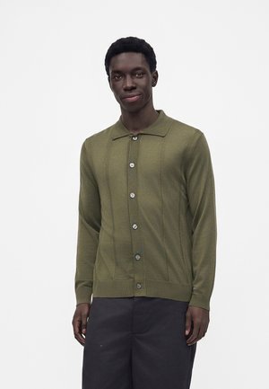 Jeune homme portant un cardigan vert olive boutonné et un pantalon noir, debout devant un fond blanc uni.