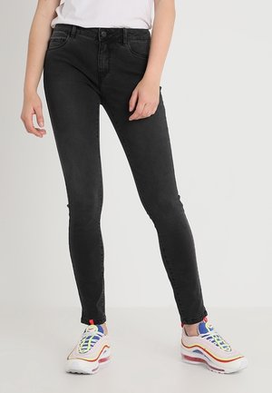 Femme portant un jean skinny noir et des baskets colorées, debout sur un fond clair uni.