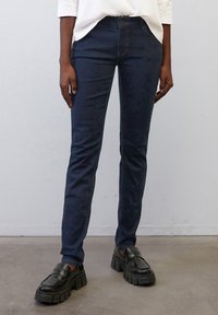 Tmavě modré, úzké džíny s klasickým designem pěti kapes a mírně elastickou texturou, spárované s robustními černými mokasíny.