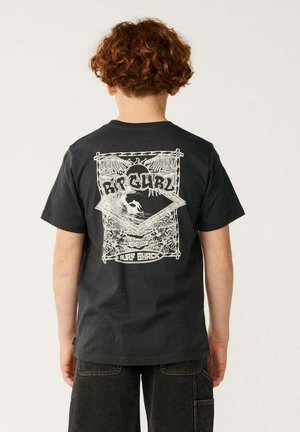 Niño con cabello rizado que lleva una camiseta negra de Rip Curl con un gráfico blanco de surfista y diseño tropical en la parte trasera.