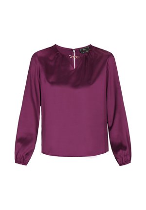 Violeta satīna blūze ar garām piedurknēm, saplacinātu kakla izgriezumu un zelta aksesuāriem priekšā. Gluda tekstūra un vienkāršs dizains.