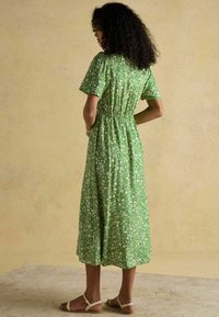 Grünes, blümchenbedrucktes Midi-Kleid mit kurzen Ärmeln, tailliertem Schnitt, fließendem Rock und leichtem Stoff. Kombiniert mit einfachen beigen Sandalen.