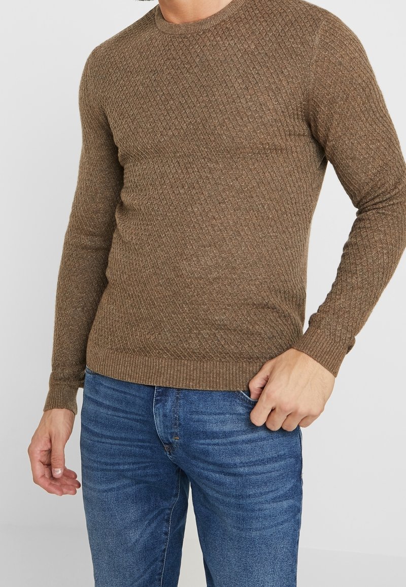 Homme portant un pull marron ajusté et texturé à manches longues et un jean bleu, debout avec une main dans la poche.