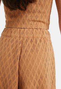 Femme portant un haut sans manches en crochet beige et un pantalon taille haute assorti avec un motif en diamant.