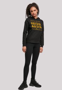 Fekete kapucnis pulóver sárga "Hakuna Matata" felirattal és geometrikus mintával. Fekete leggings-szel és fekete sportcipővel párosítva. Sima anyag, laza fazon.