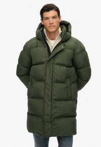 Hombre vestido con un abrigo de invierno acolchado verde oliva con capucha, sobre un suéter beige con cremallera y jeans azules, frente a un fondo blanco.