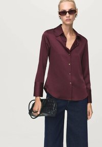 Burgundy blouse with a classic collar, long sleeves, gold buttons, and a curved hem, paired with dark wide-leg jeans and a black woven handbag.