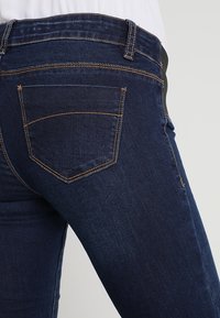 Jeans de mezclilla azul oscuro con bolsillos traseros visibles y costuras en contraste, usados por una persona vestida con una blusa blanca.