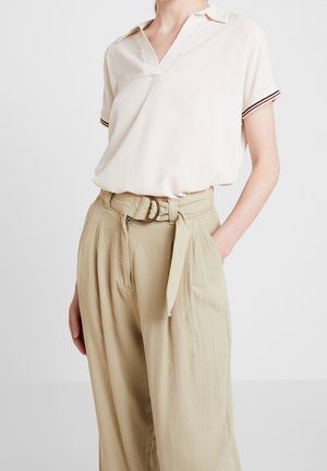 Mujer que lleva una camisa polo crema de manga corta con puños a rayas metida dentro de unos pantalones beige de tiro alto, con cinturón y pernera ancha.