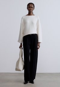 Fehér kötött pulóver széles ujjakkal, fekete, magas derekú nadrággal párosítva. Kiegészítők között világos színű válltáska található. Simaságú textúra.