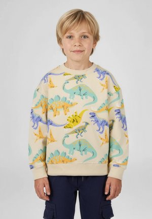 Blondýnský chlapec v krémové mikině s barevnými ilustracemi dinosaurů a tmavě modrých cargo kalhotách stojí před jednobarevným pozadím.