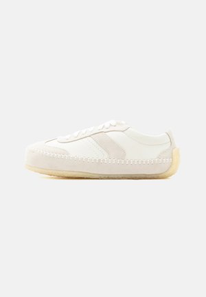 Witte lage sneaker met gestikte details, suède accenten, veterluiting en beige rubberen zool op een effen achtergrond.