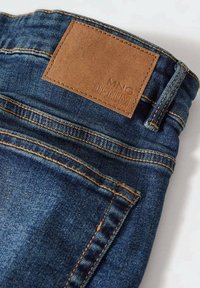 Jean en denim bleu foncé avec une patch en cuir beige portant l'inscription "MNG DENIM" au niveau de la taille, présentant des coutures visibles et des poches.