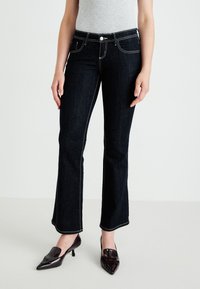 Mørk denimjeans med en tettsittende design, hvit kontrastsøm og en lett flare ved kanten, sammen med glinsende mørke sko.