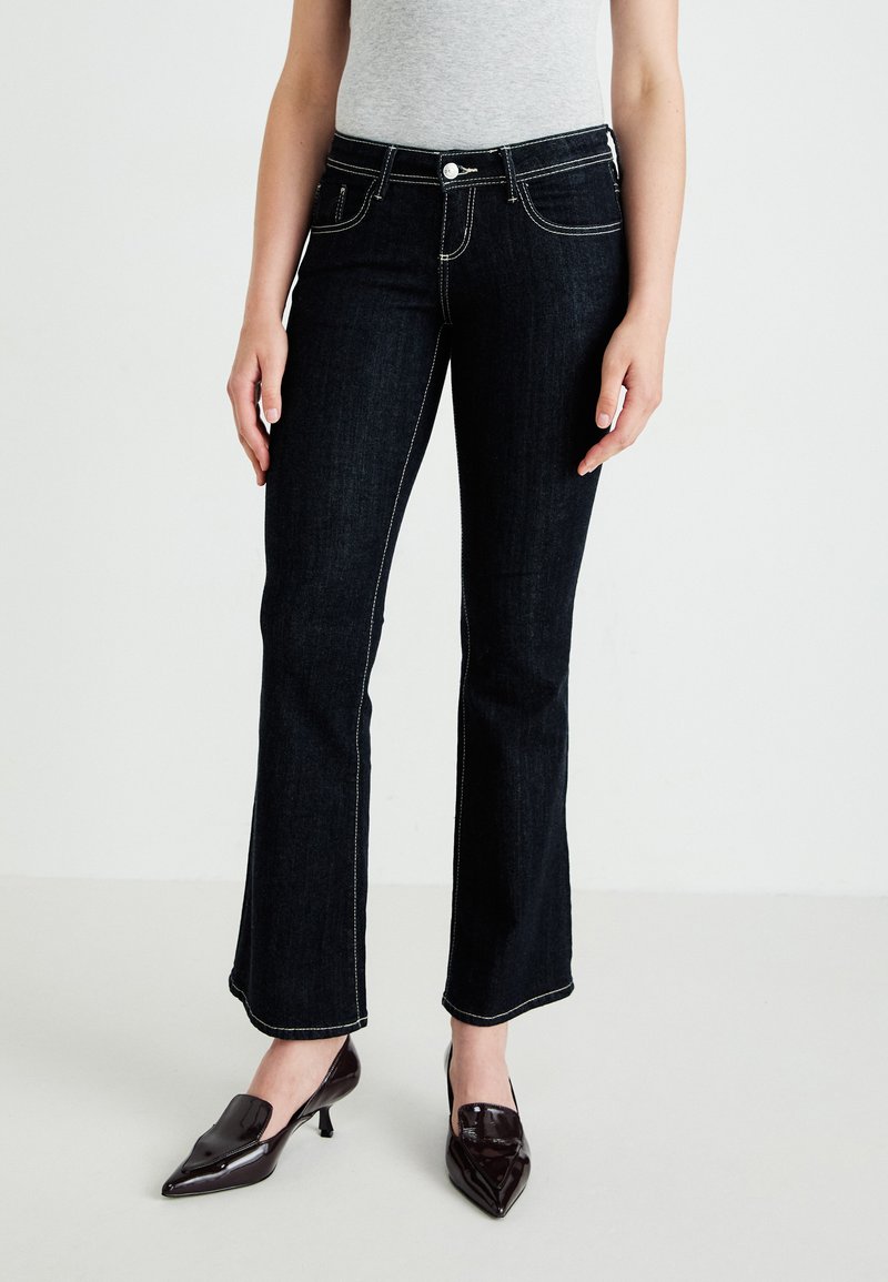 Mørk denimjeans med en tettsittende design, hvit kontrastsøm og en lett flare ved kanten, sammen med glinsende mørke sko.