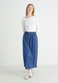 Gonna maxi in denim blu con bottoni frontali, cintura elastica e spacco laterale. Abbinata a una maglietta bianca a maniche lunghe e scarpe da ginnastica.