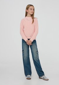 Jasnoróżowy sweter robiony na drutach z wysokim kołnierzem i teksturowanym wzorem, w połączeniu z niebieskimi spodniami cargo z bocznymi kieszeniami i luźnym krojem.