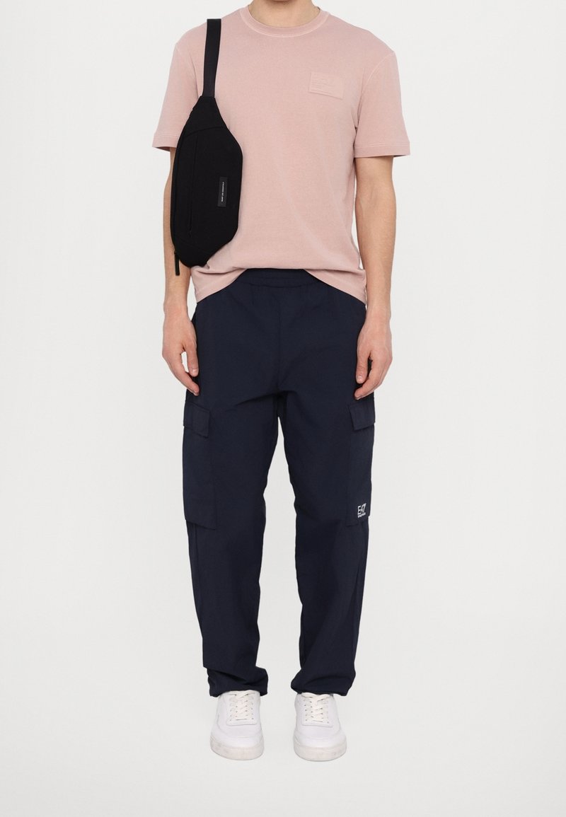 Lichtroze T-shirt met korte mouwen en een borstzakje, gecombineerd met marineblauwe cargobroek met zijzakken, en witte sneakers. Zwart schoudertasje gedragen.